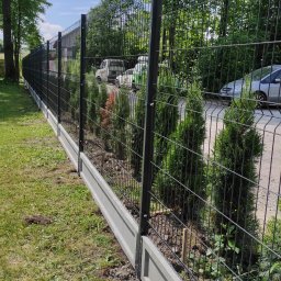 TYMACOMPANY SPÓŁKA Z OGRANICZONĄ ODPOWIEDZIALNOŚCIĄ - Długa, czarna siatka ogrodzeniowa z betonową podmurówką, otaczająca posesję z widocznymi tujami wzdłuż ogrodzenia i samochodami w tle.