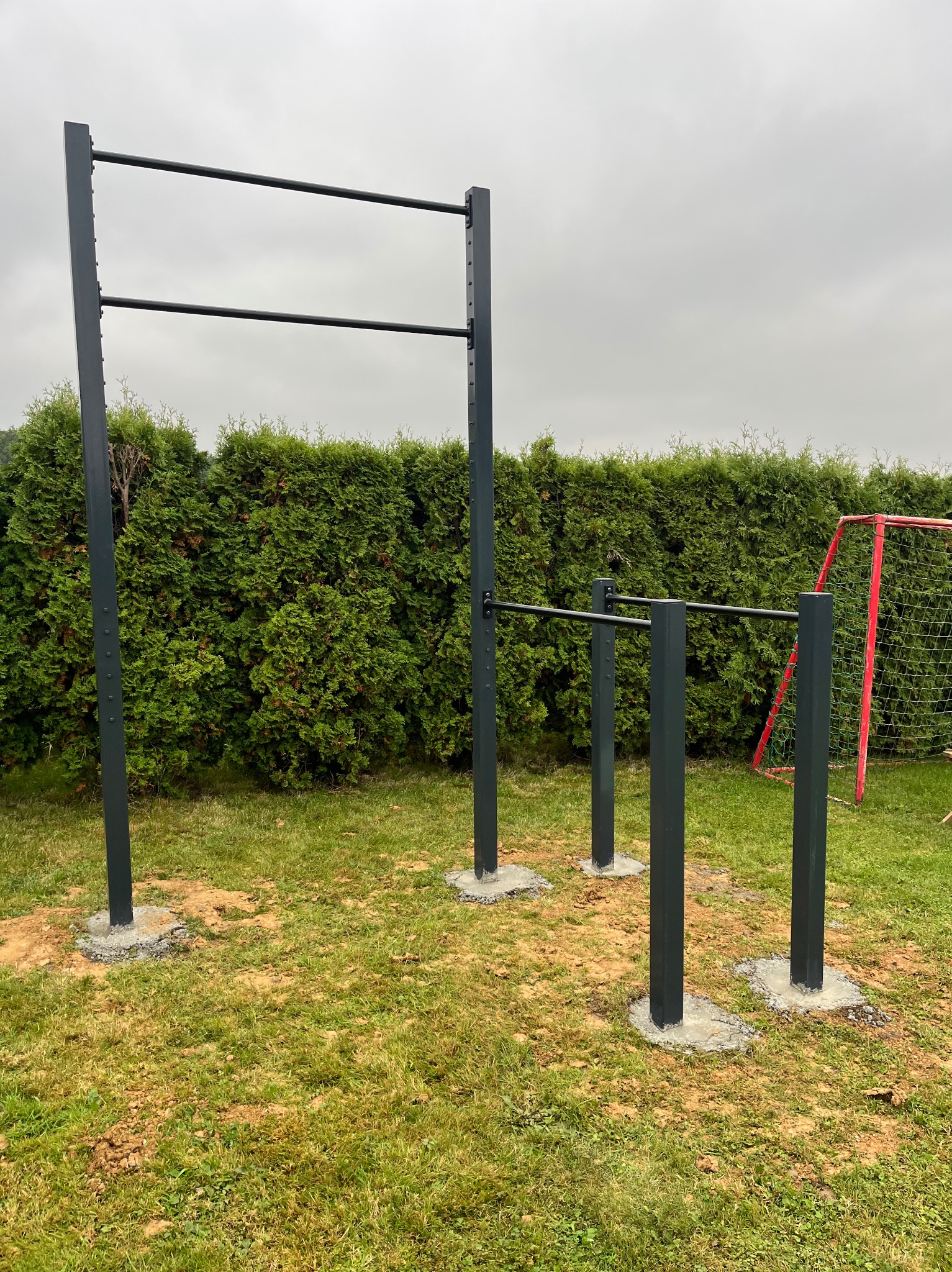 Metalowa konstrukcja do ćwiczeń na świeżym powietrzu, z drążkami na różnej wysokości, osadzona na betonowych fundamentach, na tle zielonego żywopłotu i czerwonej bramki do piłki nożnej.