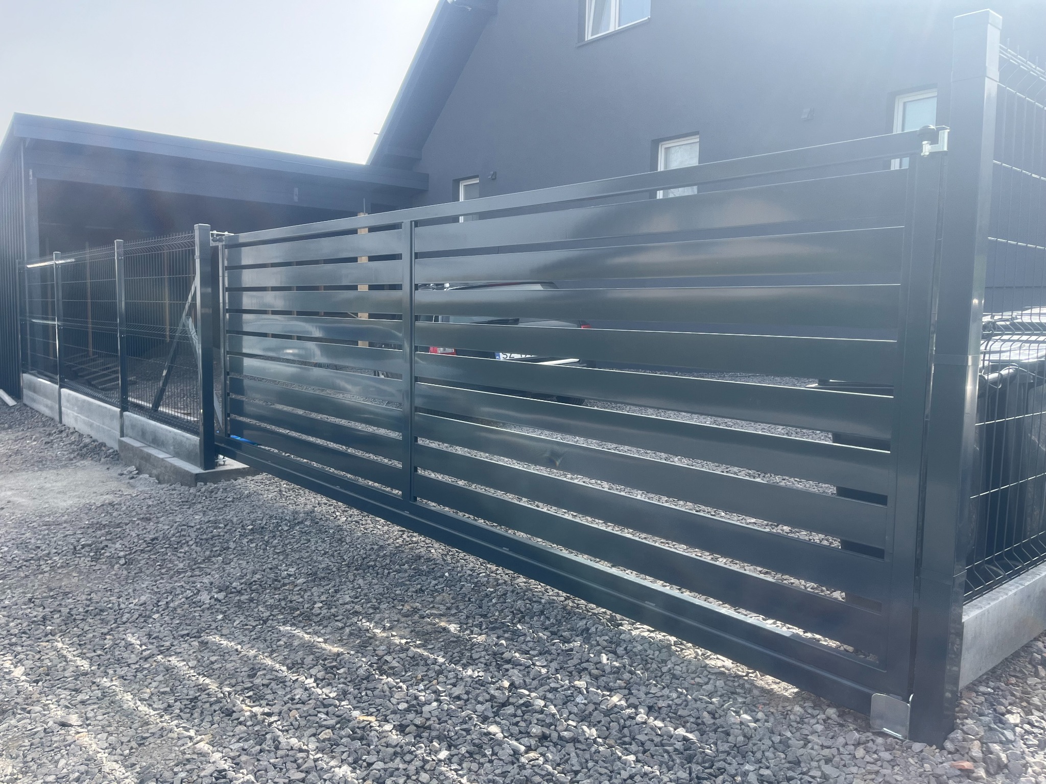 Nowoczesna, pozioma brama metalowa w kolorze antracytowym, z widoczną siatką ogrodzeniową po bokach, na tle ciemnego domu jednorodzinnego i żwirowego podjazdu.