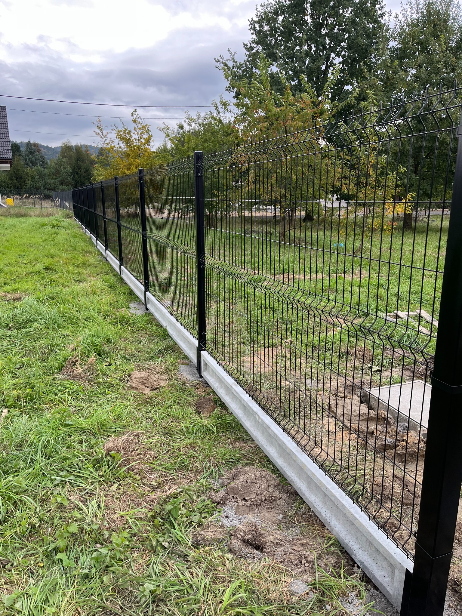 Długie, czarne ogrodzenie panelowe z podmurówką betonową na nierównym trawniku, widok z boku, pochmurne niebo.