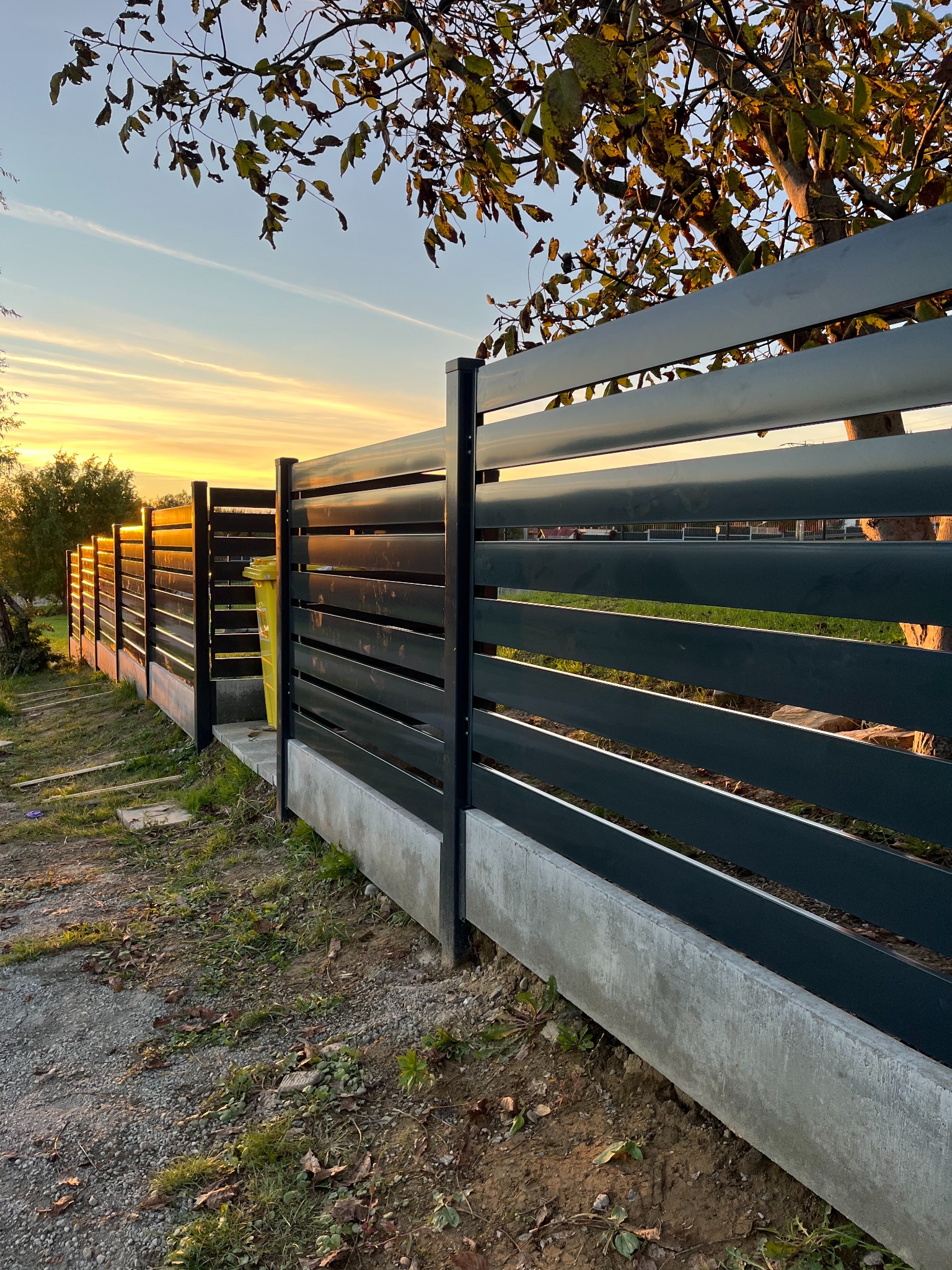 Nowoczesne ogrodzenie z poziomych, grafitowych paneli na betonowej podmurówce, widziane pod kątem o zachodzie słońca, z żółtym pojemnikiem na śmieci w tle i drzewem z opadającymi liśćmi...