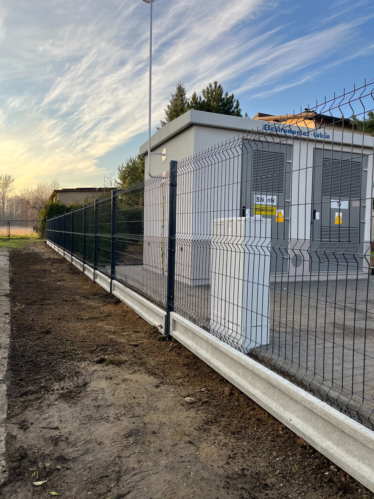 Nowoczesne ogrodzenie panelowe w kolorze grafitowym z podmurówką betonową, chroniące transformator Elektromontaż-Lublin, tło: błękitne niebo z pierzastymi chmurami.
