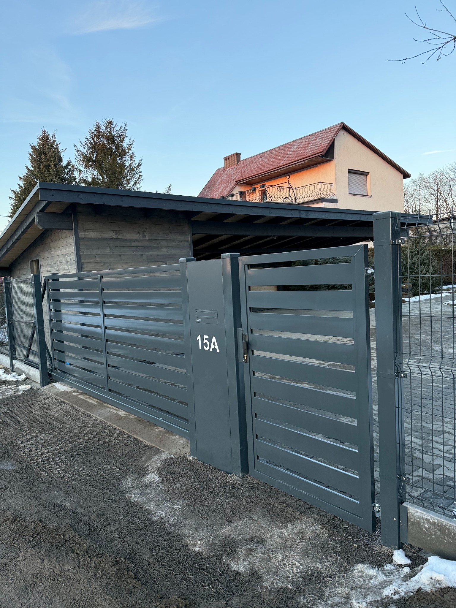 Nowoczesna, przesuwna, metalowa brama w kolorze grafitowym z poziomymi panelami, numer domu 15A, widok z zewnątrz na tle domu z czerwonym dachem i zieleni, częściowo ośnieżony podjazd.