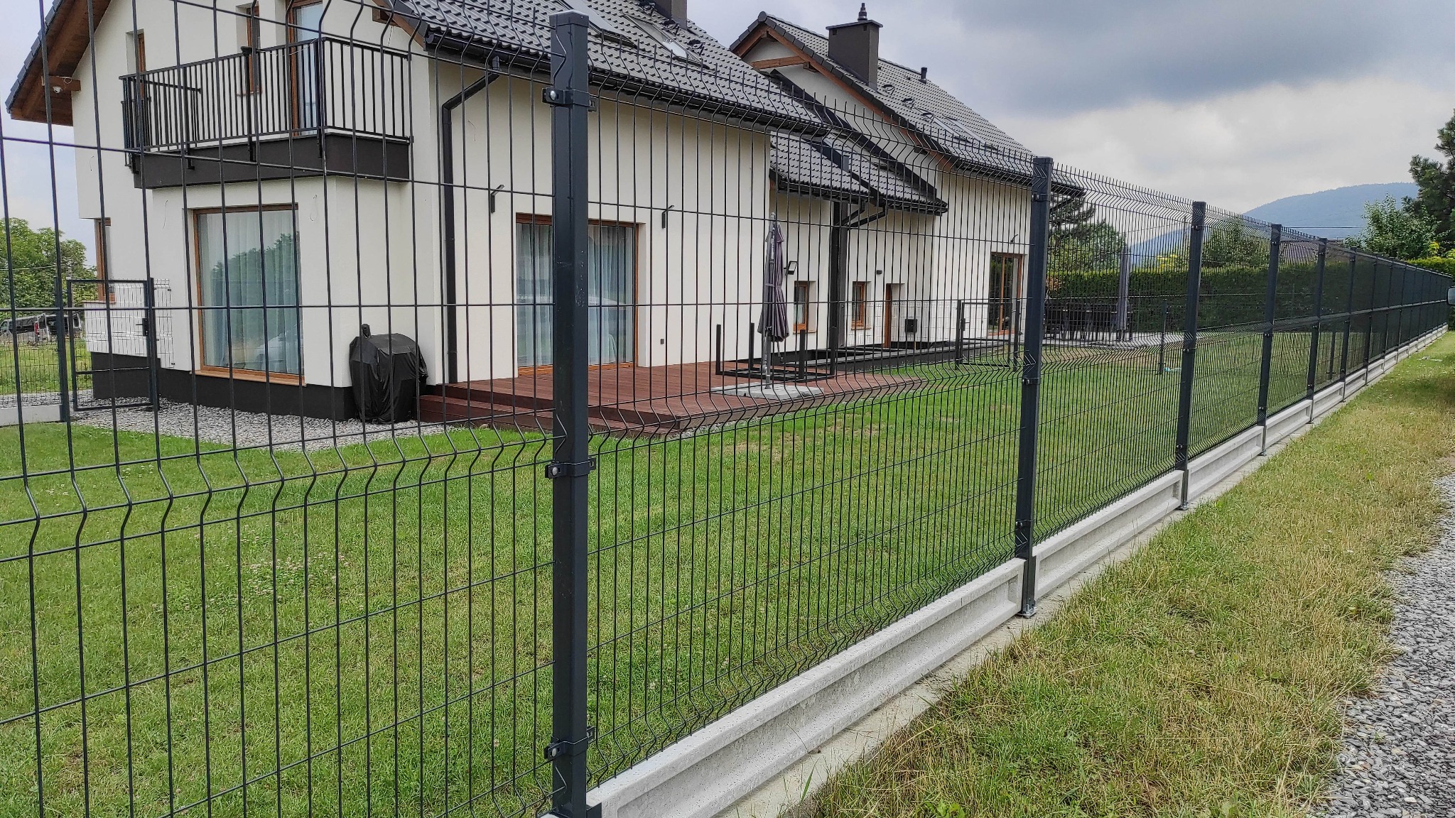 Nowoczesne ogrodzenie panelowe z podmurówką betonową otaczające dom z drewnianym tarasem i balkonem, widok od strony drogi.