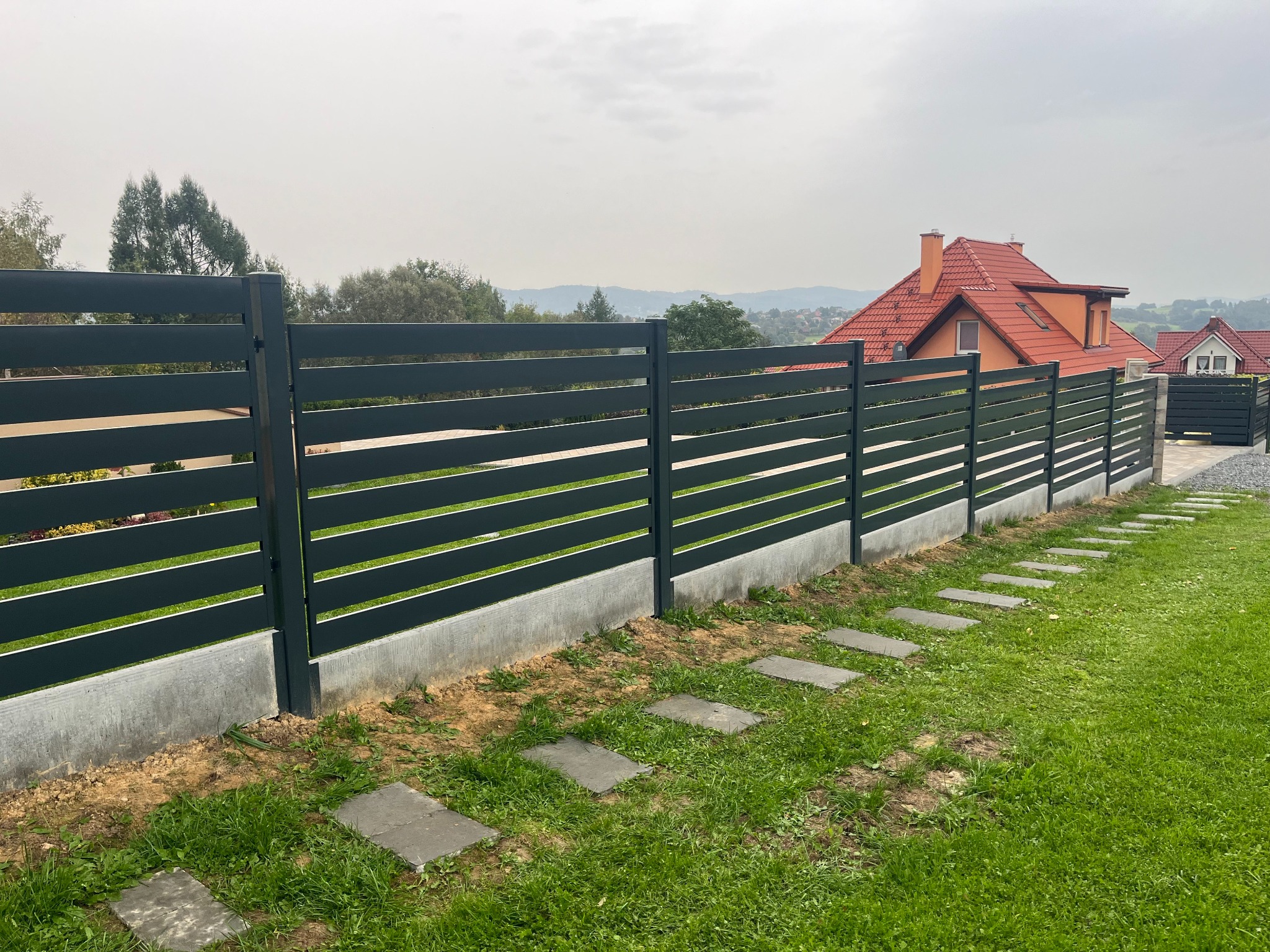 Nowoczesne, poziome ogrodzenie panelowe w kolorze grafitowym, osadzone na betonowej podmurówce. W tle widoczne domy z czerwonymi dachami i zieleń drzew na wzgórzach pod pochmurnym niebem. Ścieżka...