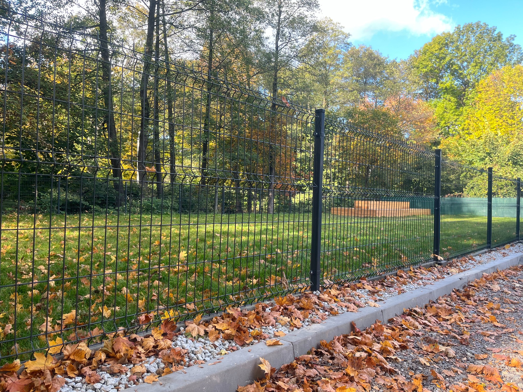 Nowoczesne ogrodzenie panelowe w kolorze grafitowym, z widocznymi słupkami, zamontowane na obrzeżu trawnika, z opadłymi liśćmi jesiennymi przy krawężniku, w tle drzewa w jesiennej kolorystyce.