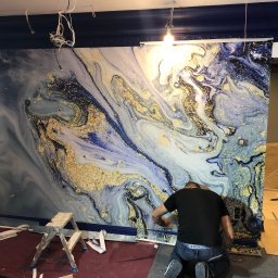 QUALI WNĘTRZA SPÓŁKA Z OGRANICZONĄ ODPOWIEDZIALNOŚCIĄ - Montaż tapety z efektem marmuru i złotych żył w pomieszczeniu z ciemnoniebieską listwą przysufitową, widoczne drabiny i narzędzia.