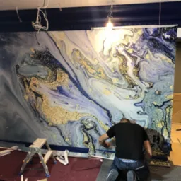 Montaż tapety z efektem marmuru i złotych żył w pomieszczeniu z ciemnoniebieską listwą przysufitową, widoczne drabiny i narzędzia.