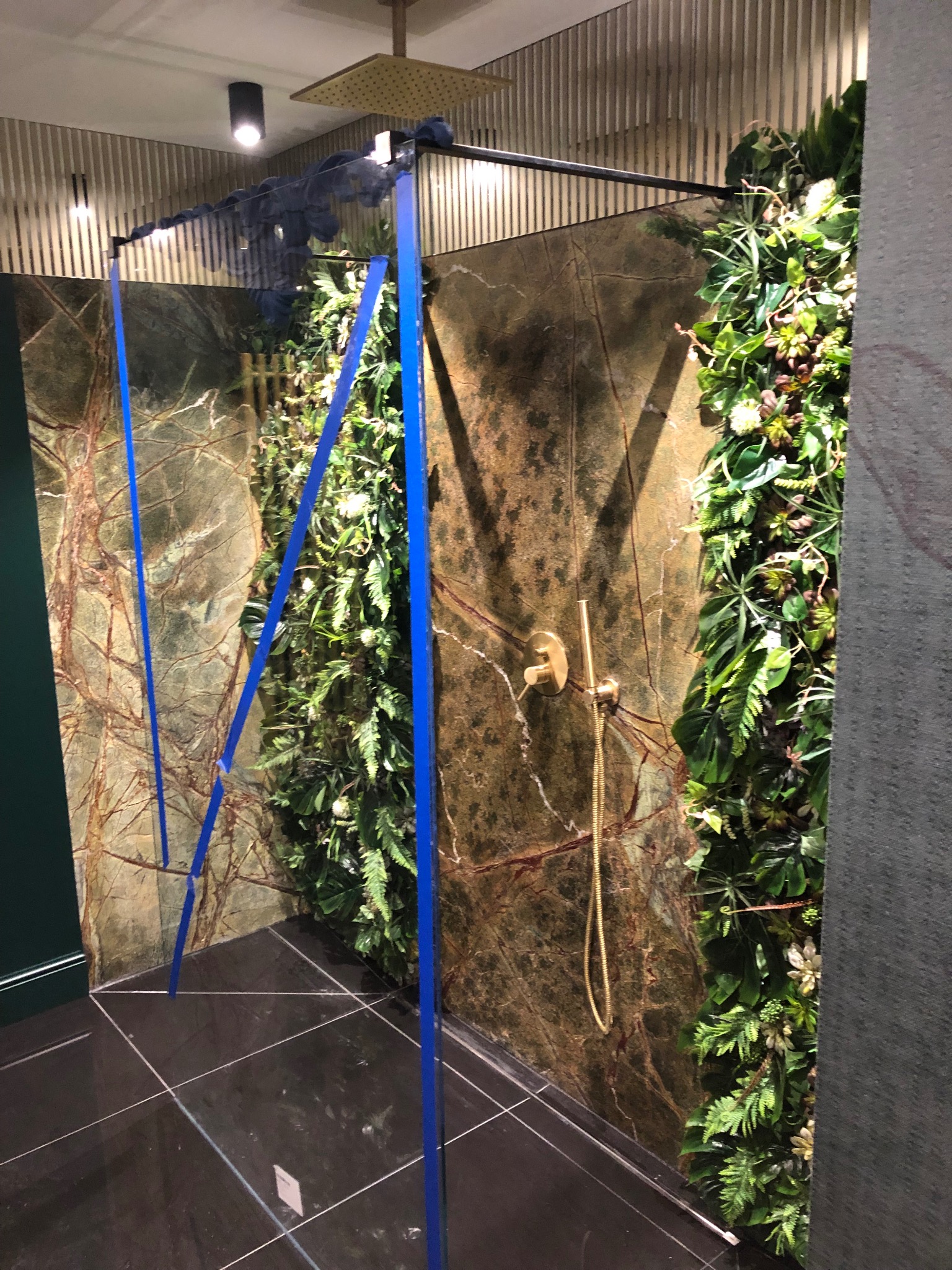 Nowoczesna kabina prysznicowa z elementami zieleni, marmurowymi płytami i złotą armaturą w trakcie montażu, podparta niebieskimi stemplami.