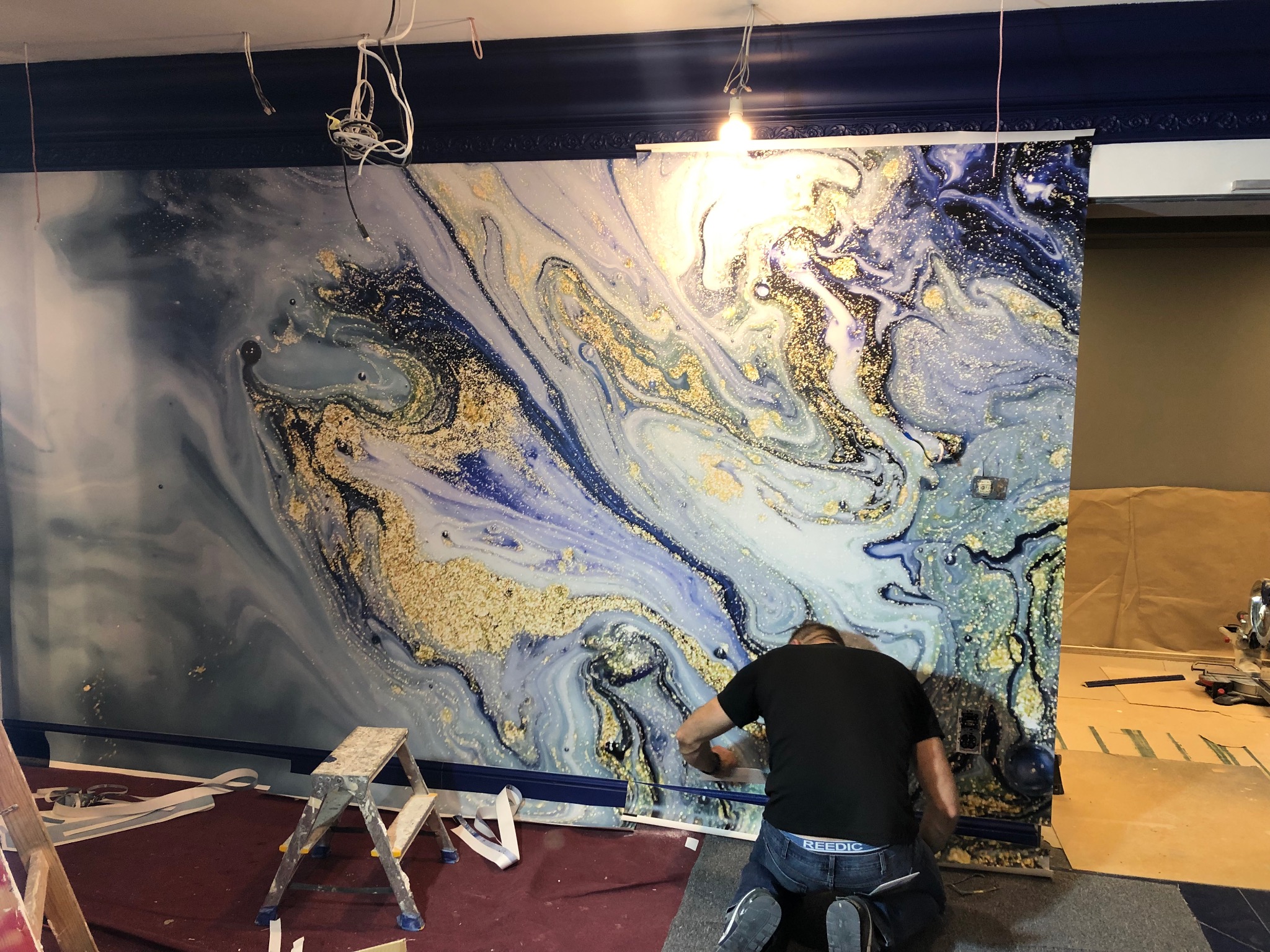 Montaż tapety z efektem marmuru i złotych żył w pomieszczeniu z ciemnoniebieską listwą przysufitową, widoczne drabiny i narzędzia.