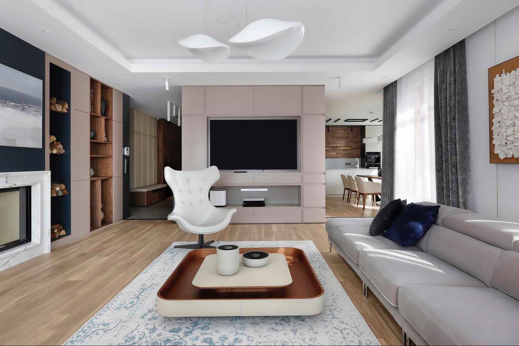 Elegancki salon w Chwaszczynie z jasną podłogą, szarą sofą z granatowymi poduszkami, białym fotelem obrotowym, zabudową RTV w odcieniach beżu i marmurowym kominkiem z drewnem.