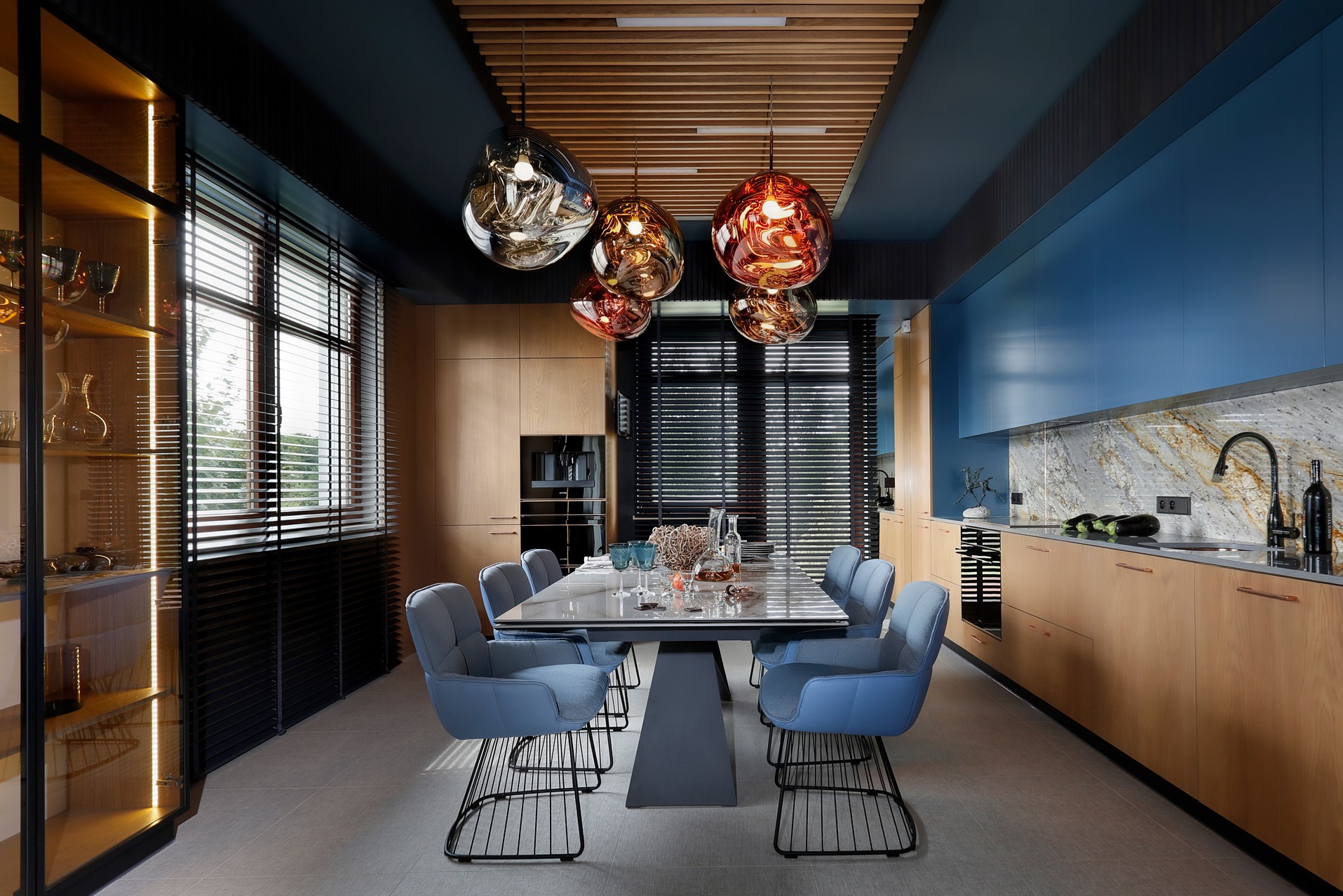 Nowoczesna jadalnia z otwartą kuchnią: niebieskie szafki górne, drewniane szafki dolne, marmurowy backsplash, designerskie lampy wiszące nad stołem z krzesłami w odcieniach błękitu.