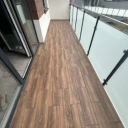 Długi balkon z podłogą wyłożoną brązowymi płytkami imitującymi drewno, z widokiem na nowoczesne budynki mieszkalne i flagę. Szklane balustrady z metalowymi ramami.