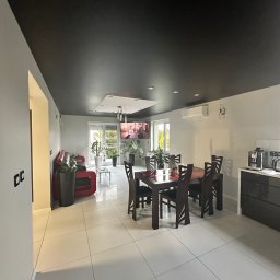 PerfectFinish - Przestronne wnętrze z czarnym sufitem podwieszanym, białymi ścianami i podłogą z płytek, z widoczną częścią jadalną z ciemnym drewnianym stołem i krzesłami oraz częścią wypoczynkową z czerwoną sofą.