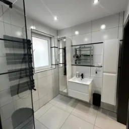 Nowoczesna łazienka w bieli i czerni z kabiną prysznicową ze szklaną ścianką, grzejnikiem drabinkowym, umywalką z szafką i lustrem z podświetleniem LED.