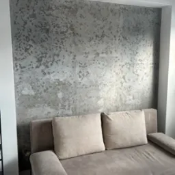 Ściana z dekoracyjną, szarą fakturą imitującą kamień, widoczna nad beżową sofą z poduszkami w neutralnych barwach.