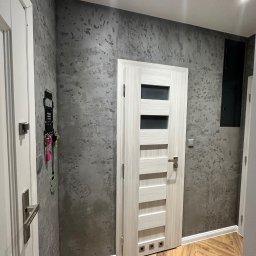 PerfectFinish - Wnętrze przedpokoju z szarą ścianą o fakturze betonu, jasnymi drzwiami z przeszkleniami i drewnianą podłogą ułożoną w jodełkę, widoczne akcesoria na ścianie i oświetlenie sufitowe.