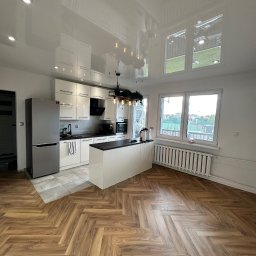 PerfectFinish - Jasna kuchnia z wyspą, parkietem w jodełkę i błyszczącym, odbijającym światło sufitem napinanym, widok na okna z widokiem na boisko.