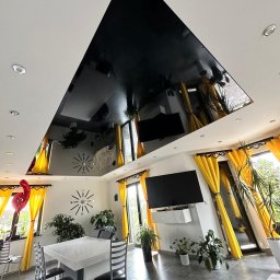 PerfectFinish - Salon z czarnym, lustrzanym sufitem napinanym, odbijającym wyposażenie: stół, krzesła, rośliny i żółte zasłony.