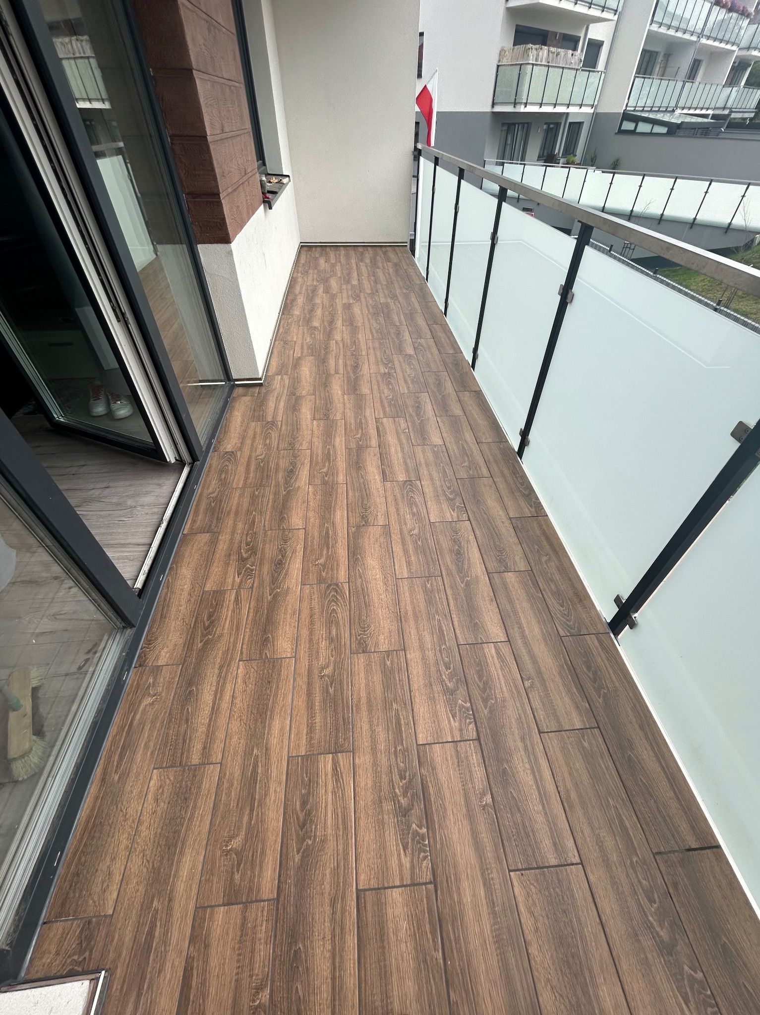 Długi balkon z podłogą wyłożoną brązowymi płytkami imitującymi drewno, z widokiem na nowoczesne budynki mieszkalne i flagę. Szklane balustrady z metalowymi ramami.