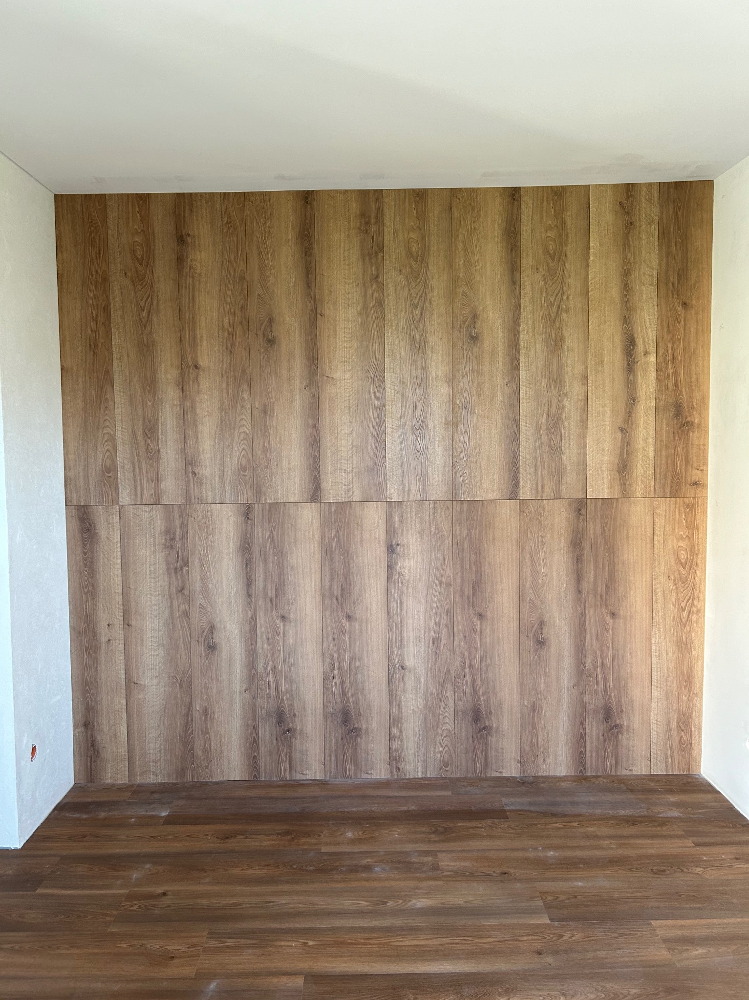 Ściana i podłoga wykończone panelami imitującymi drewno, widoczne łączenie paneli na ścianie, surowy stan ścian bocznych.