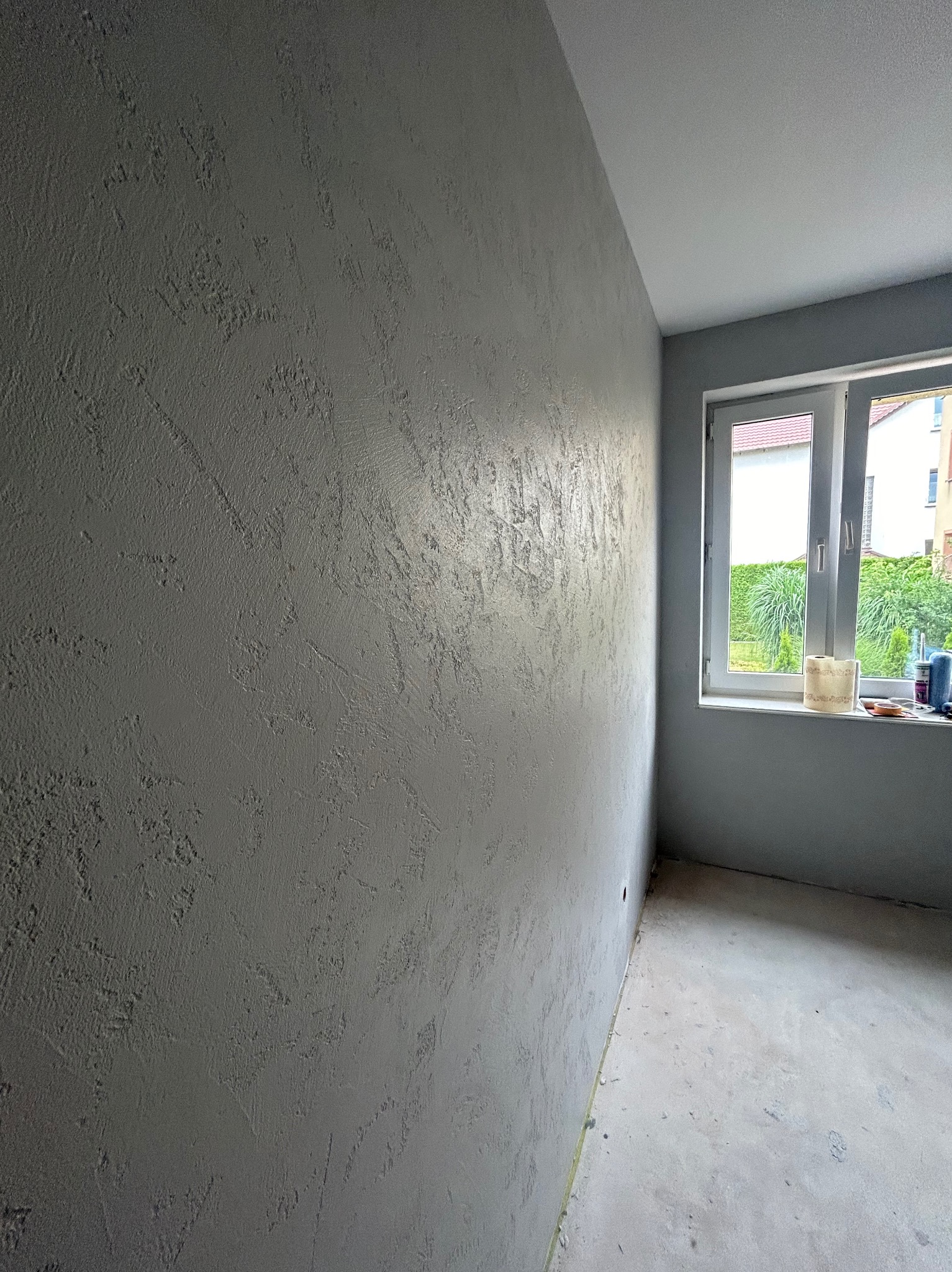 Szara ściana z dekoracyjną fakturą w pokoju, widok na okno z roślinnością na zewnątrz, widoczne akcesoria malarskie na parapecie.