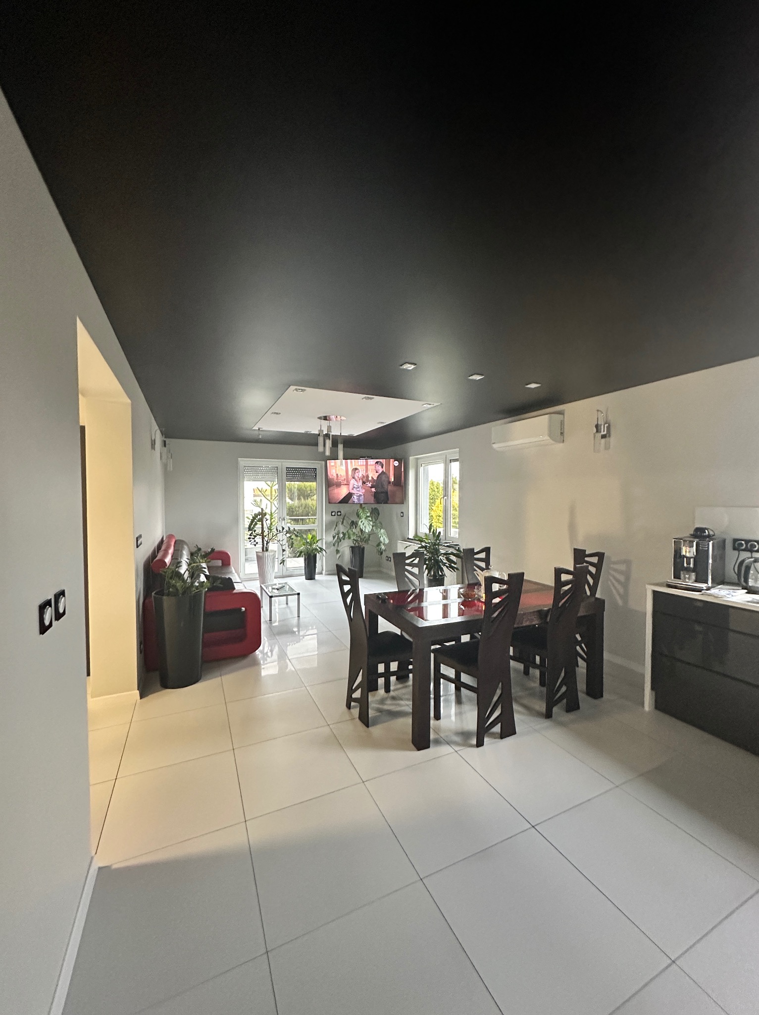 Przestronne wnętrze z czarnym sufitem podwieszanym, białymi ścianami i podłogą z płytek, z widoczną częścią jadalną z ciemnym drewnianym stołem i krzesłami oraz częścią wypoczynkową z czerwoną sofą.