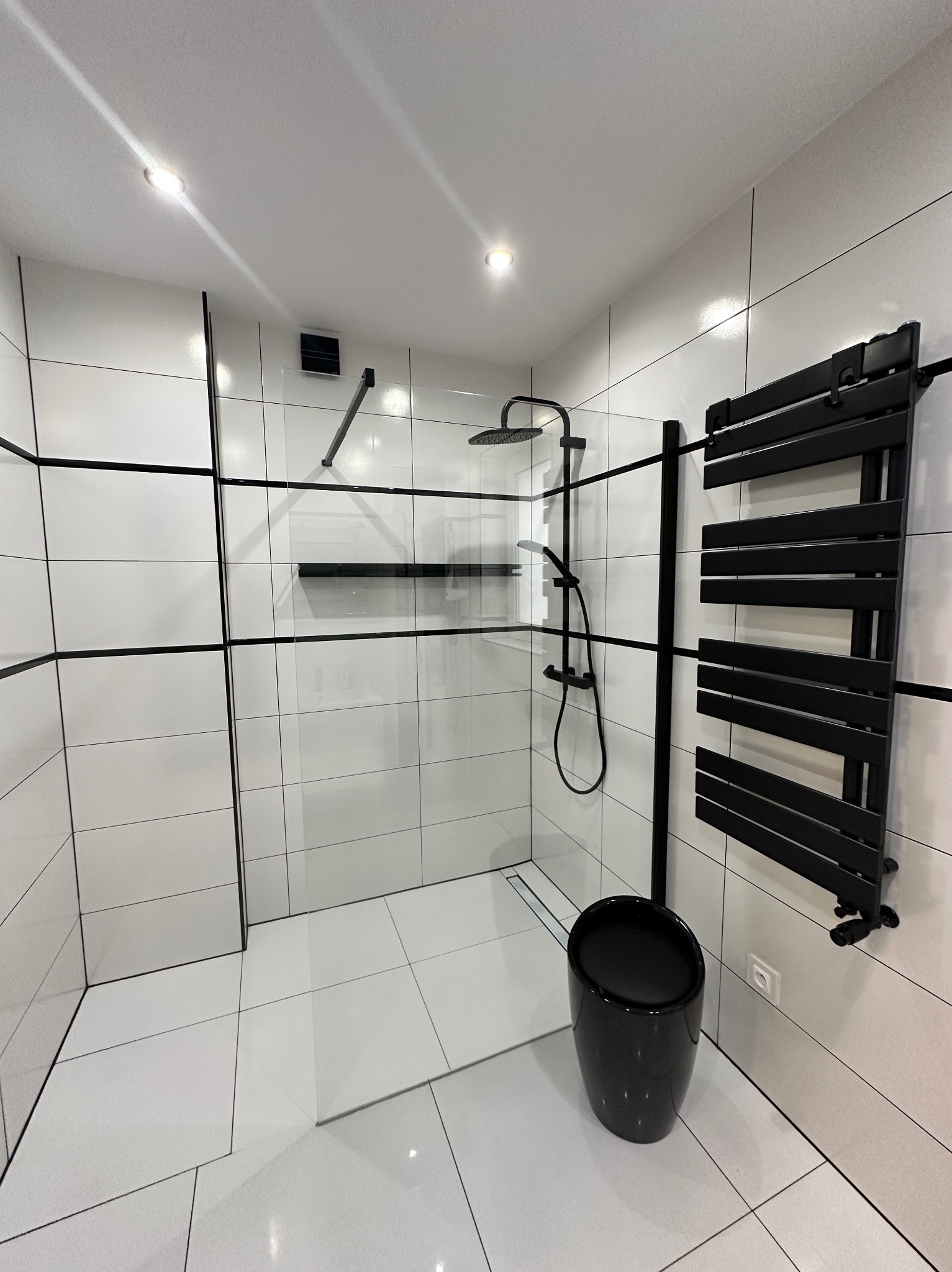 Nowoczesna łazienka z kabiną prysznicową typu walk-in, wykończona białymi płytkami z czarnymi fugami, czarną armaturą i grzejnikiem drabinkowym w tym samym kolorze, oraz czarnym, błyszczącym stołkiem.