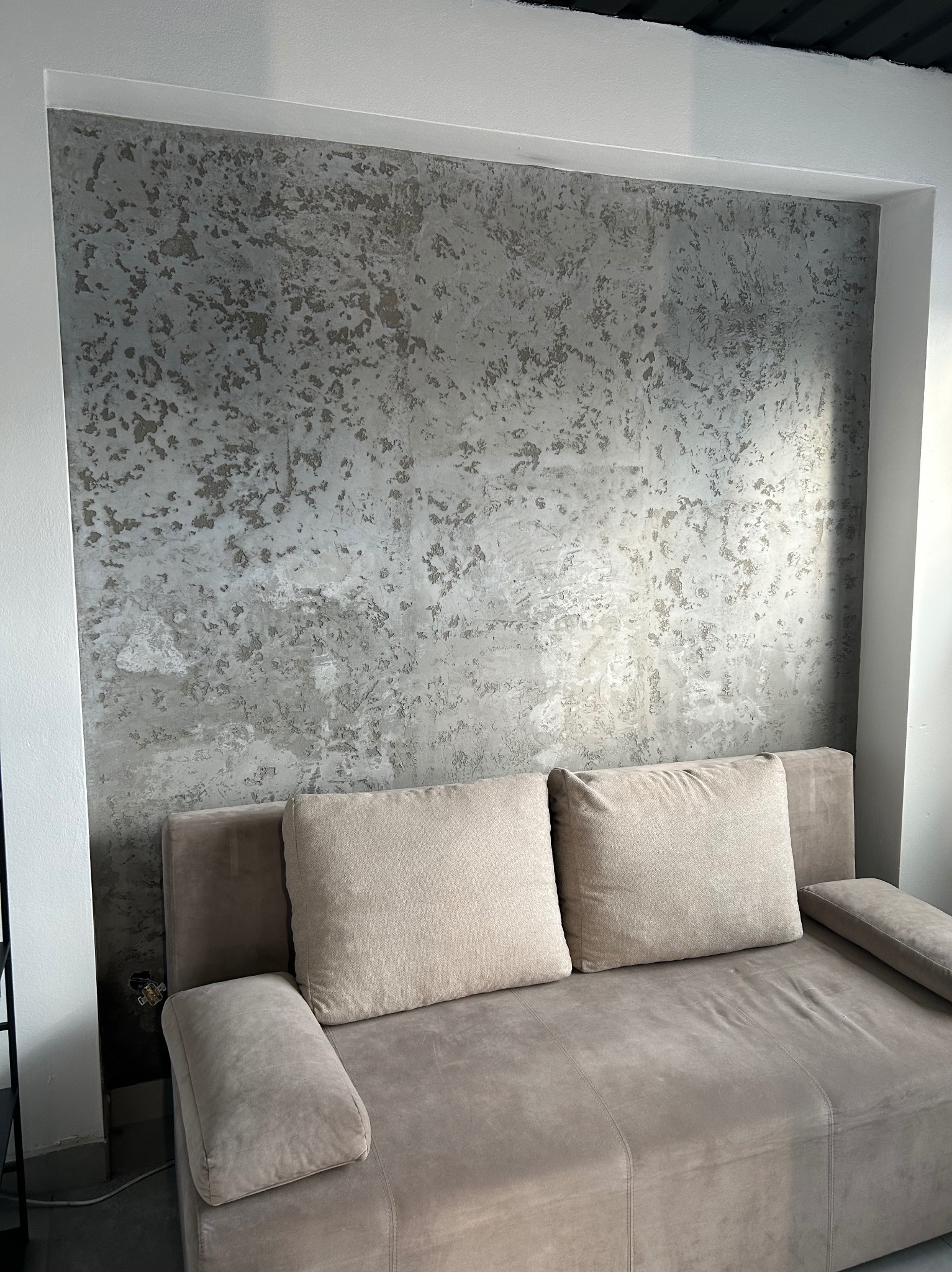 Ściana z dekoracyjną, szarą fakturą imitującą kamień, widoczna nad beżową sofą z poduszkami w neutralnych barwach.