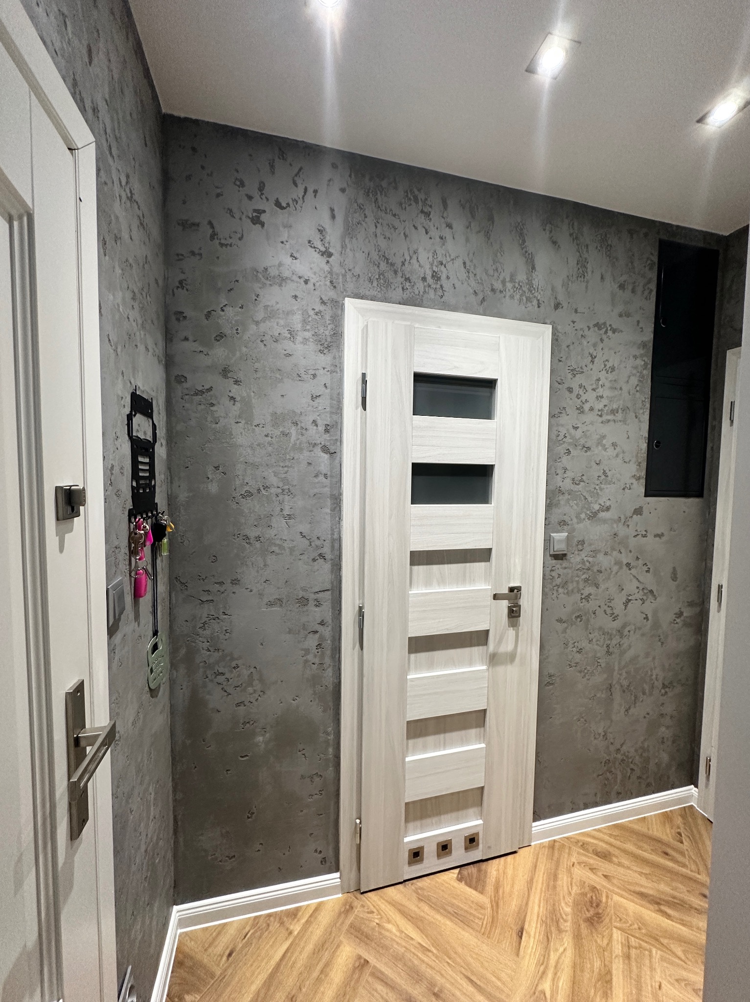 Wnętrze przedpokoju z szarą ścianą o fakturze betonu, jasnymi drzwiami z przeszkleniami i drewnianą podłogą ułożoną w jodełkę, widoczne akcesoria na ścianie i oświetlenie sufitowe.