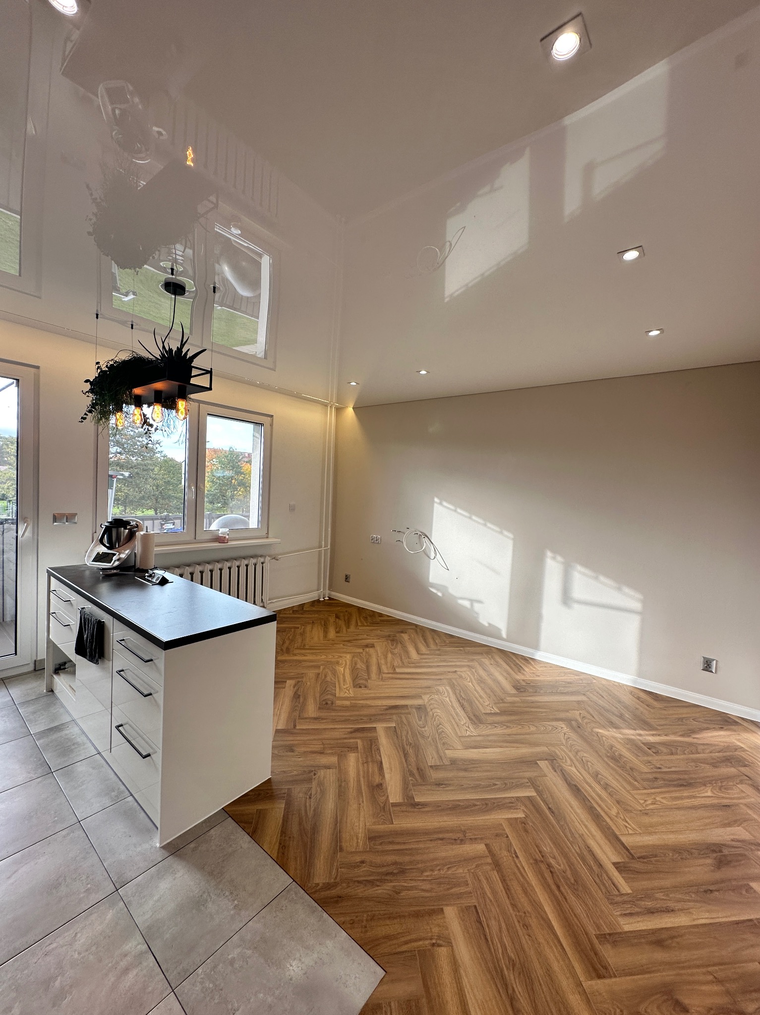 Nowoczesne wnętrze po remoncie: parkiet ułożony w jodełkę, biała wyspa kuchenna z czarnym blatem, błyszczący sufit napinany z oświetleniem punktowym i wisząca lampa z roślinami.