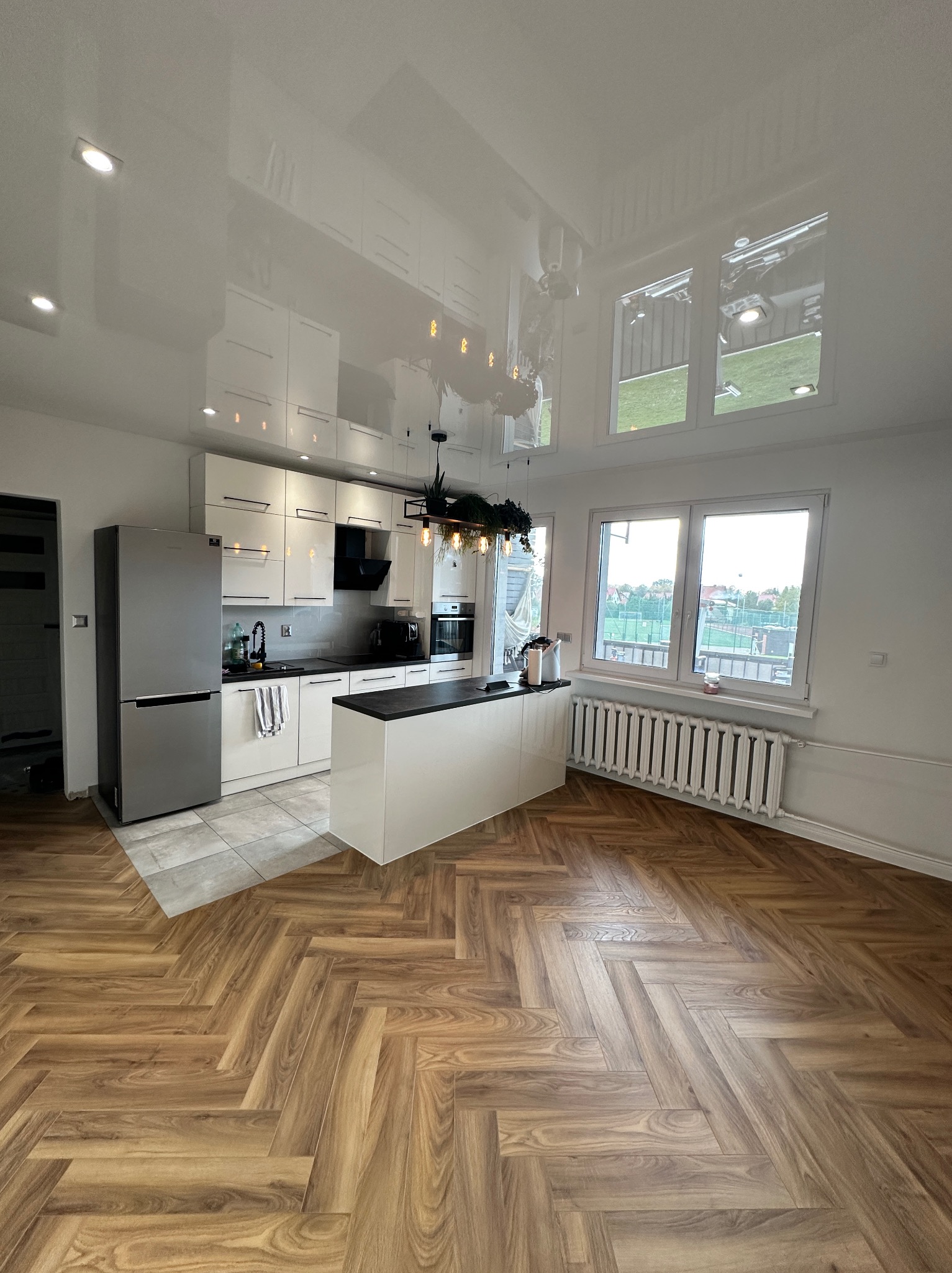 Jasna kuchnia z wyspą, parkietem w jodełkę i błyszczącym, odbijającym światło sufitem napinanym, widok na okna z widokiem na boisko.