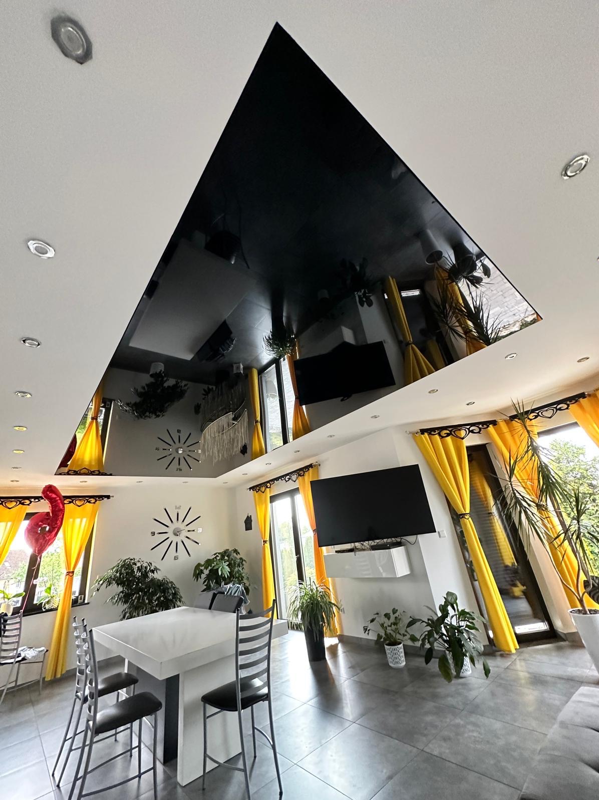Salon z czarnym, lustrzanym sufitem napinanym, odbijającym wyposażenie: stół, krzesła, rośliny i żółte zasłony.