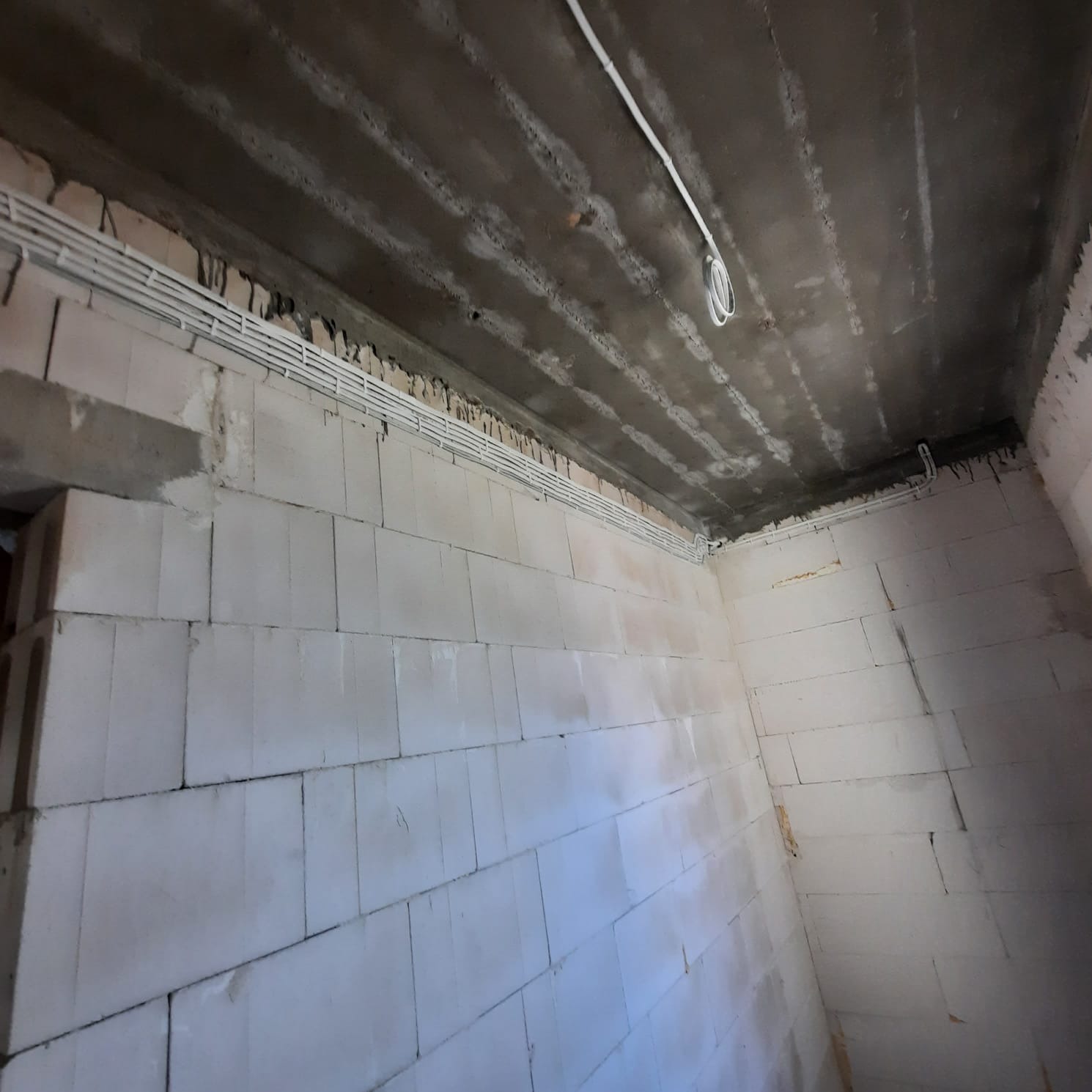 Wnętrze w trakcie remontu z widocznymi przewodami elektrycznymi ułożonymi w korytkach na ścianach i suficie z betonu.