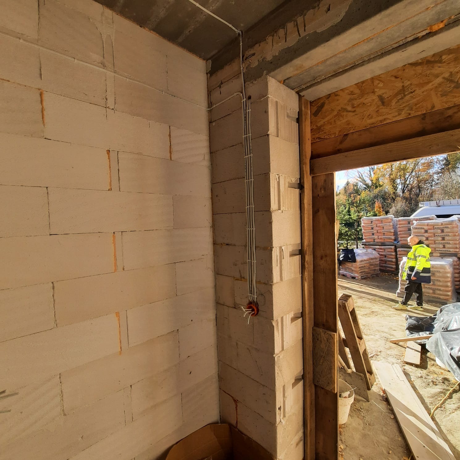 Wewnętrzny róg budynku w trakcie budowy z widocznymi przewodami elektrycznymi w białych peszlach, wychodzącymi z czerwonej puszki instalacyjnej. W tle widoczny pracownik w żółtej kamizelce i worki...