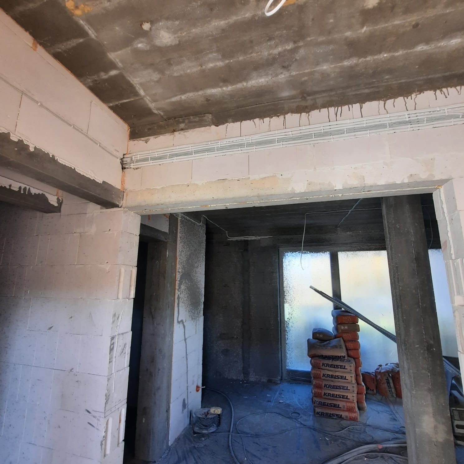 Wnętrze w trakcie budowy: surowy betonowy strop z widocznymi przewodami w białych korytkach, ściany z bloczków, worki z zaprawą Kreisel ustawione pod oknem, podłoga zabezpieczona folią.
