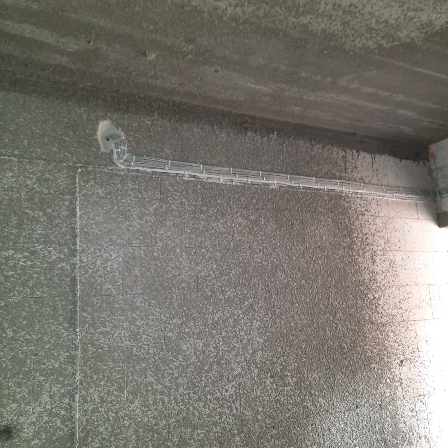 Szara ściana z betonowych bloczków z ułożonymi wzdłuż kablami elektrycznymi w białych peszlach, w rogu widoczny otwór w ścianie z wyprowadzonymi kablami.