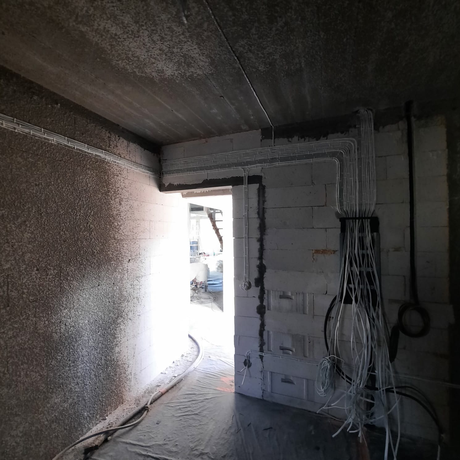 Surowy stan wnętrza w trakcie prac elektrycznych: widoczne kable prowadzone w korytkach na ścianach z betonu komórkowego i suficie, zwisające przewody z puszki, zarys drzwi w tle.