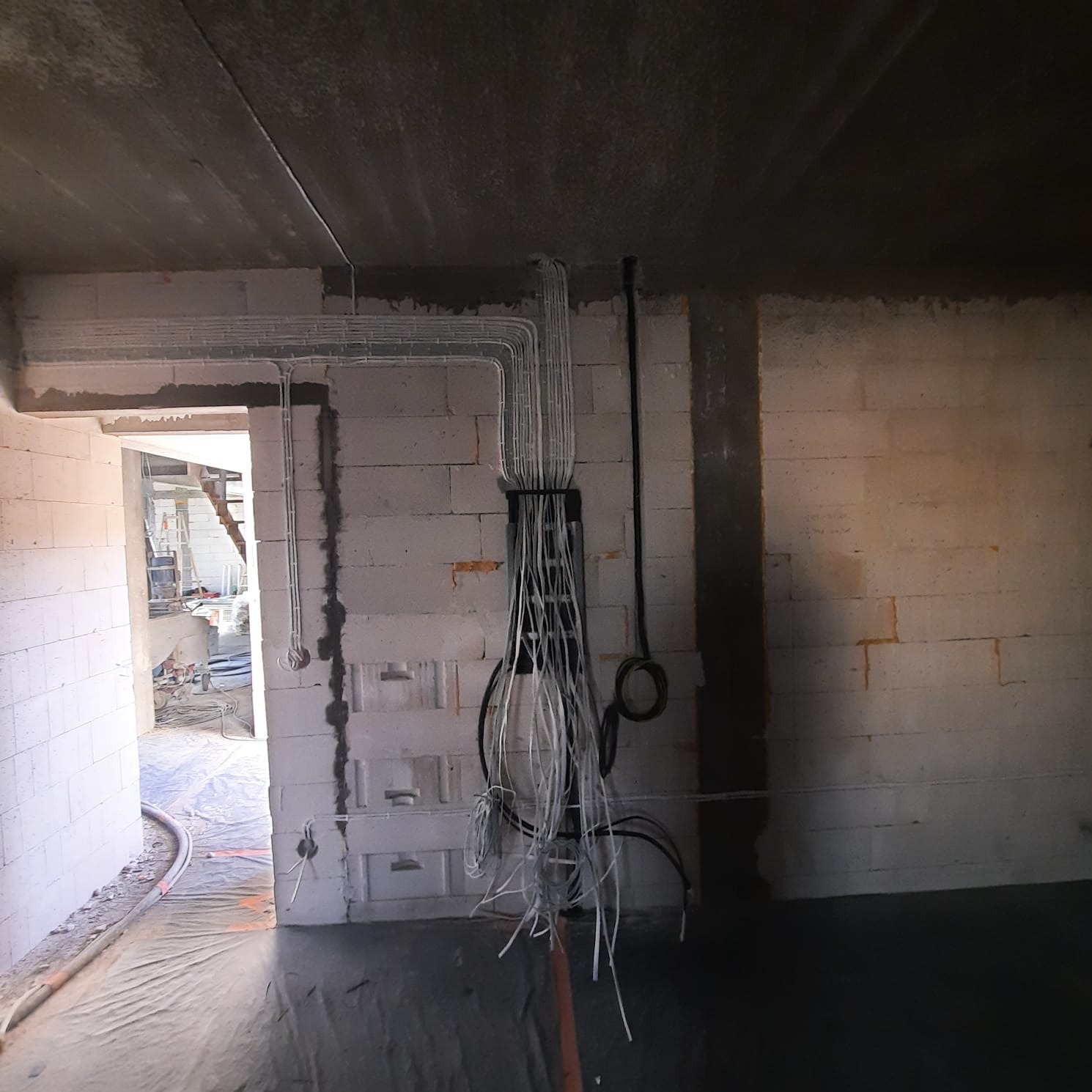 Surowy stan budynku z widoczną puszką instalacyjną i licznymi przewodami elektrycznymi, częściowo ułożonymi w korytkach na ścianie z bloczków betonowych, widok na korytarz w tle.