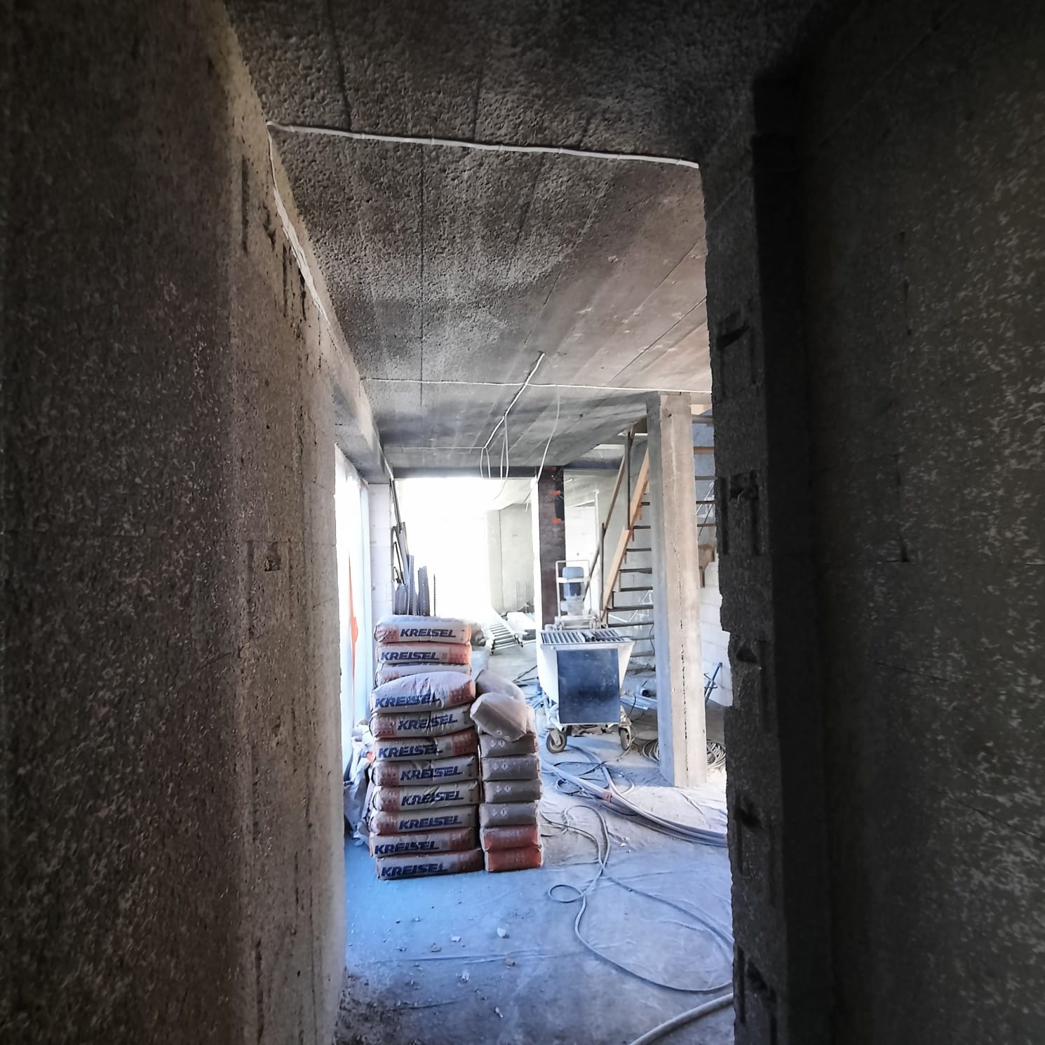 Surowy stan budynku w trakcie prac wykończeniowych: widok przez otwór w ścianie na stertę worków z zaprawą Kreisel, w tle wózek budowlany i schody, widoczne przewody elektryczne pod sufitem.