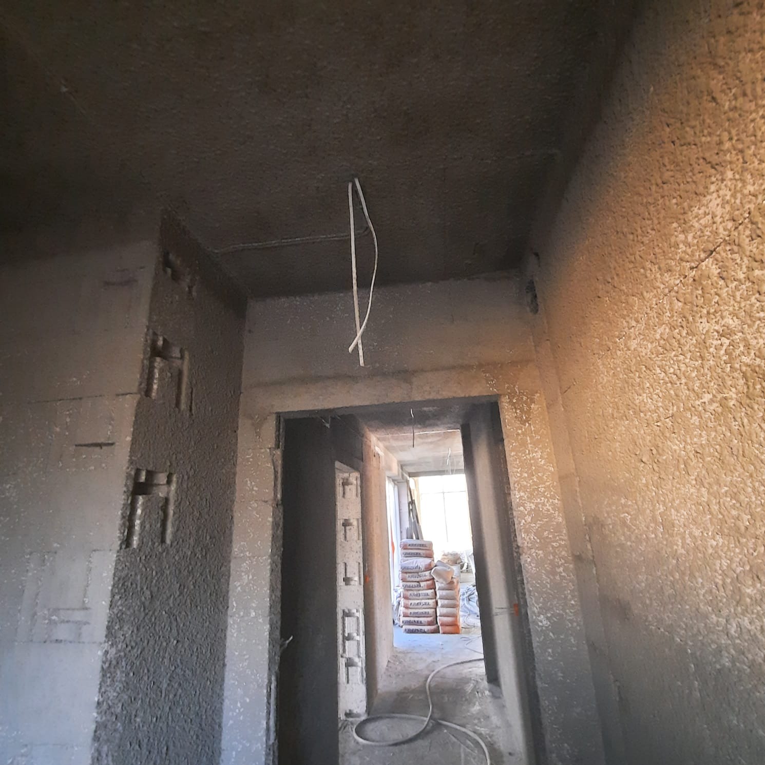 Surowy stan wnętrza w trakcie prac wykończeniowych; widoczne ściany po tynkowaniu, wystające przewody elektryczne i stosy worków z zaprawą w głębi pomieszczenia.