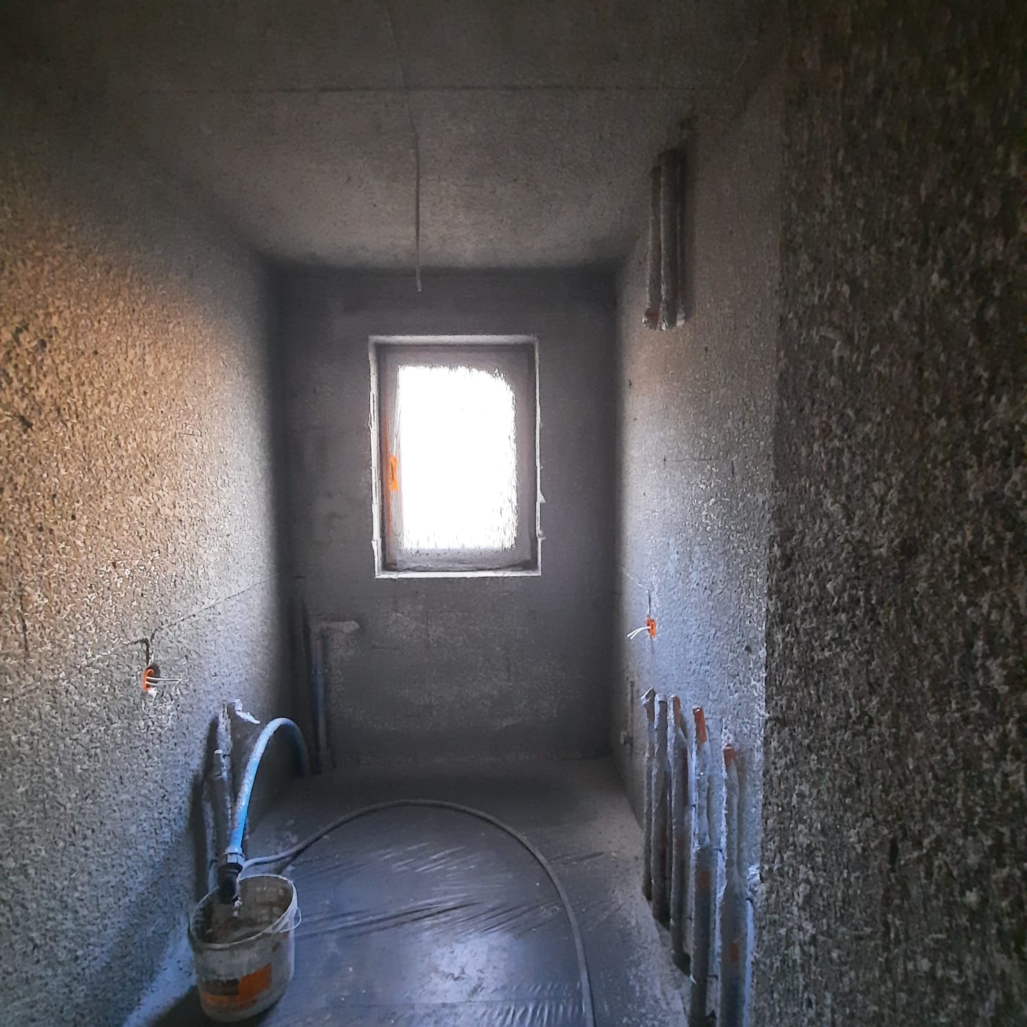 Surowy stan wnętrza z widocznymi przewodami elektrycznymi wystającymi ze ściany, przygotowanego do dalszych prac wykończeniowych, z oknem w tle i prowizorycznym oświetleniem.