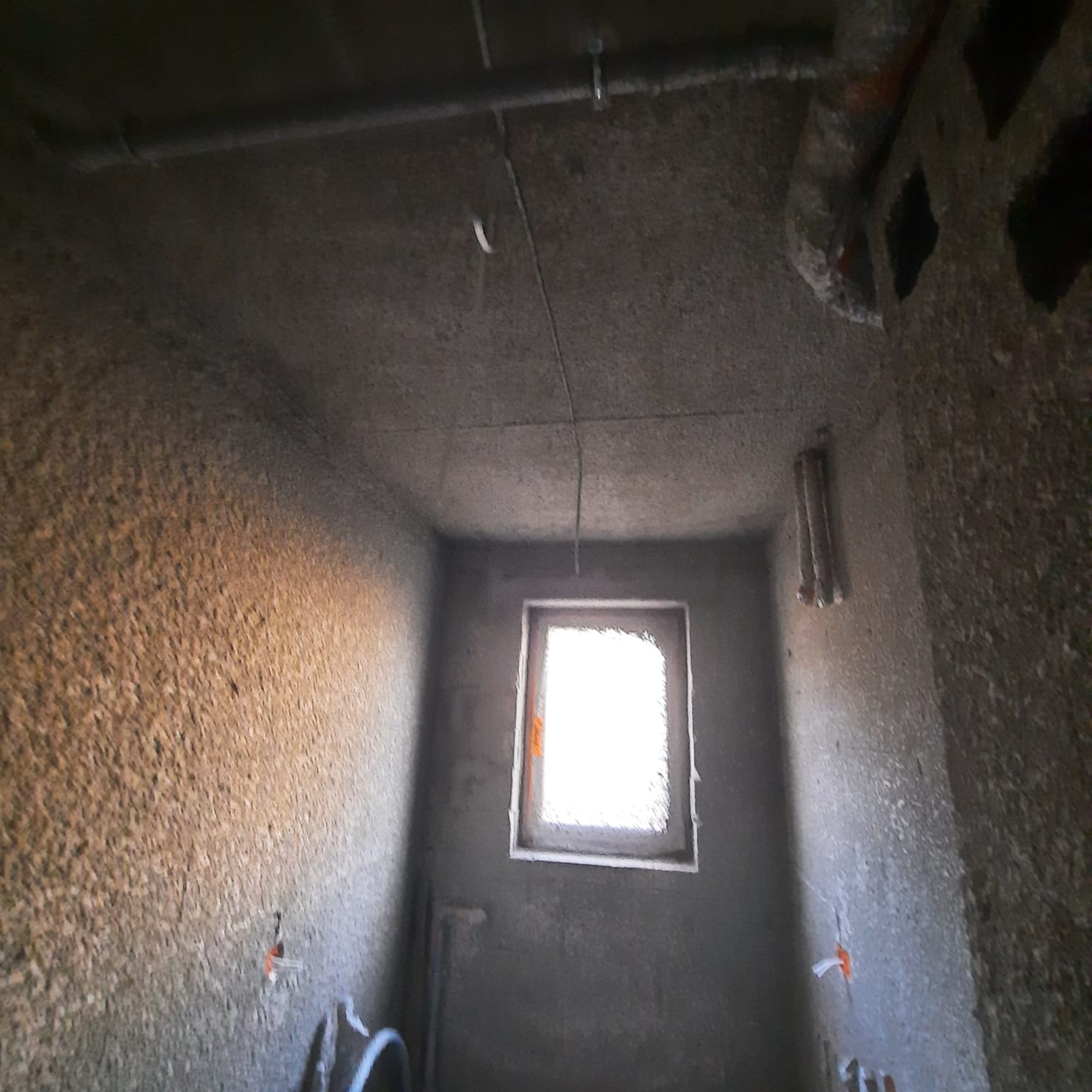 Surowe wnętrze pomieszczenia w stanie deweloperskim z widocznymi przewodami elektrycznymi wystającymi ze ścian, rurami pod sufitem i oknem z widocznym światłem dziennym.