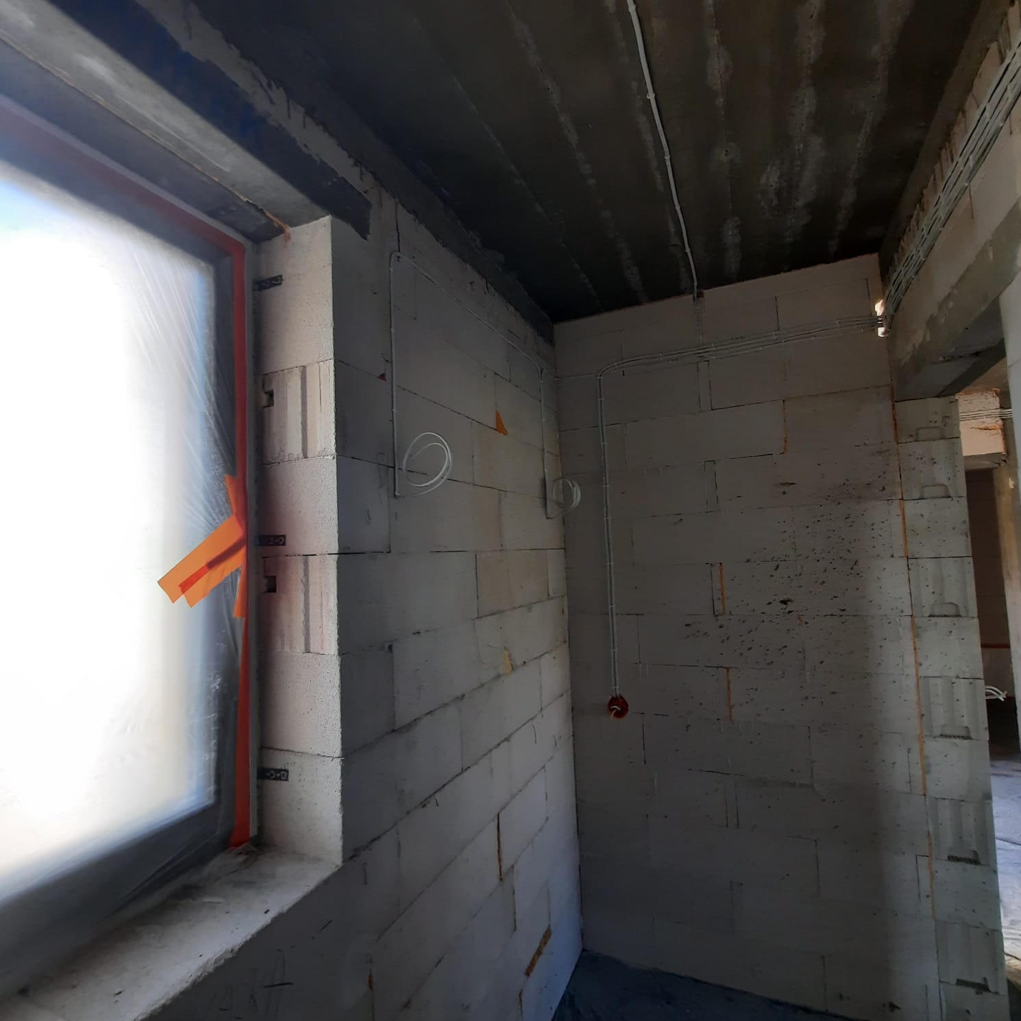 Wnętrze w stanie surowym: ściany z betonu komórkowego, okno zabezpieczone folią i taśmą, widoczne przewody elektryczne pod sufitem i na ścianie, puszka elektryczna.