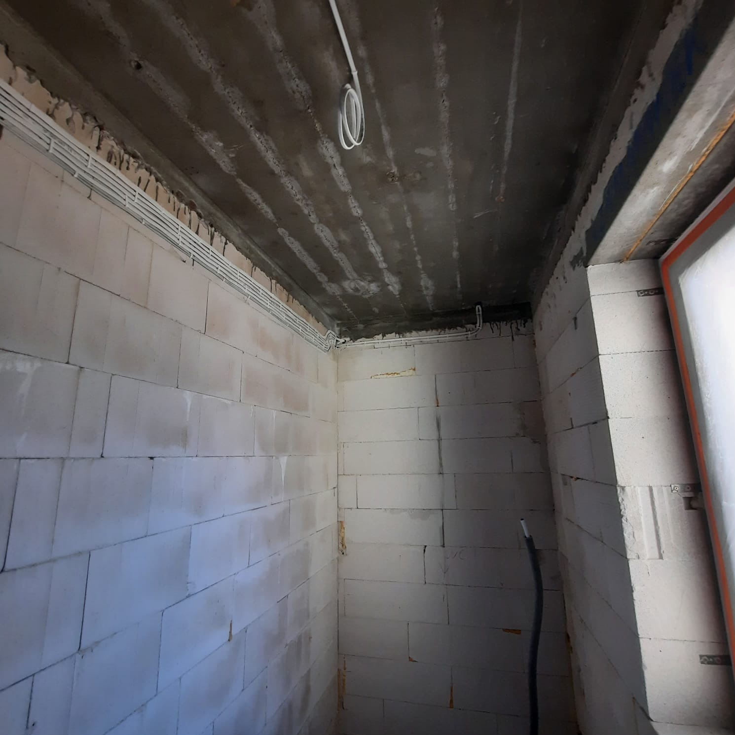 Surowy pokój z betonu komórkowego w trakcie prac wykończeniowych, widoczne białe przewody elektryczne ułożone w korytkach na suficie i ścianach, oraz czarny kabel zwisający w rogu.