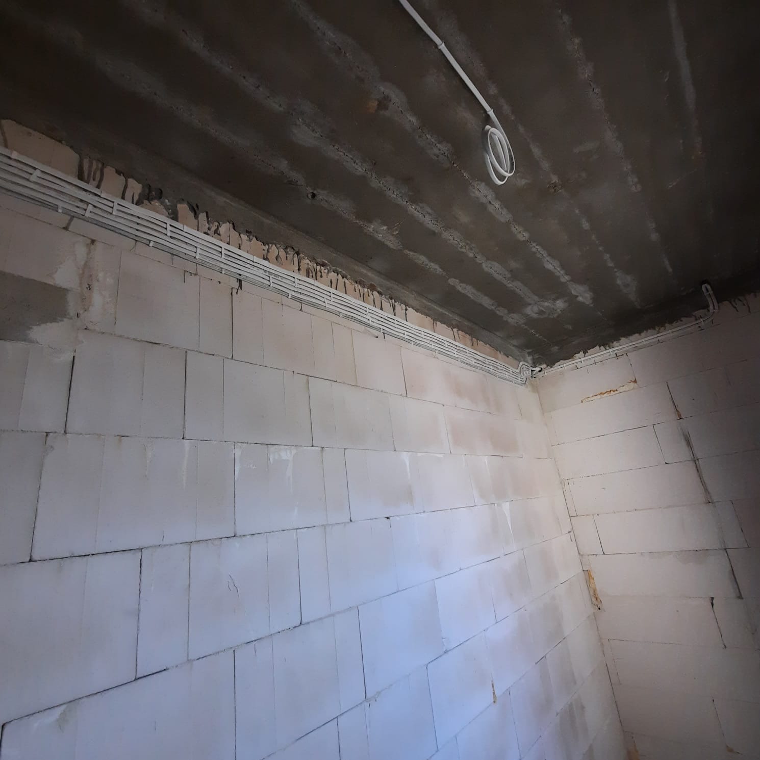 Surowy stan wnętrza z białych bloczków, z widocznymi przewodami elektrycznymi w białych listwach biegnącymi wzdłuż ściany pod betonowym sufitem. Część przewodów zwisa w pętli.
