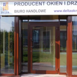 Brązowe drzwi wejściowe z przeszkleniami, widoczne z zewnątrz, z logo producenta okien i drzwi na elewacji z szarej cegły.