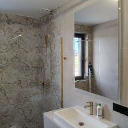 KAMIL NIEDZIELSKI KN-BUD - Elegancka łazienka z marmurowymi płytkami, złotą armaturą i dużym lustrem. Widoczna kabina prysznicowa i umywalka nablatowa. Nowoczesny design z dbałością o detale.