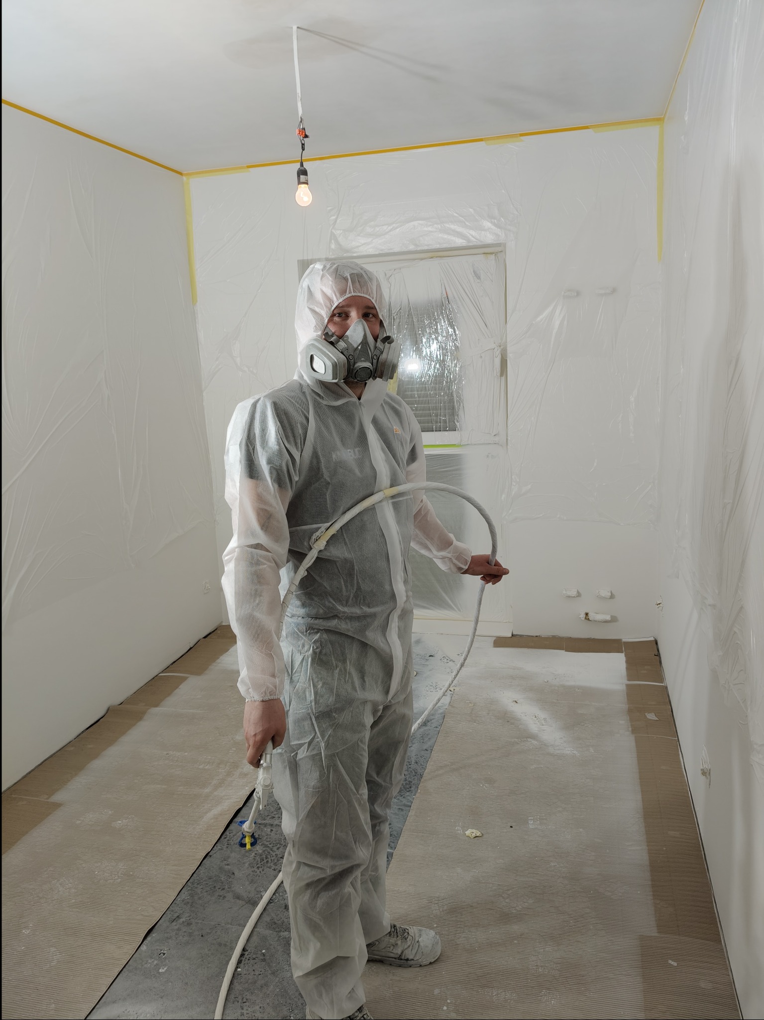 Pracownik w kombinezonie i masce maluje ściany natryskowo w pomieszczeniu zabezpieczonym folią i taśmą malarską. Widoczne narzędzie pracy i oświetlenie.