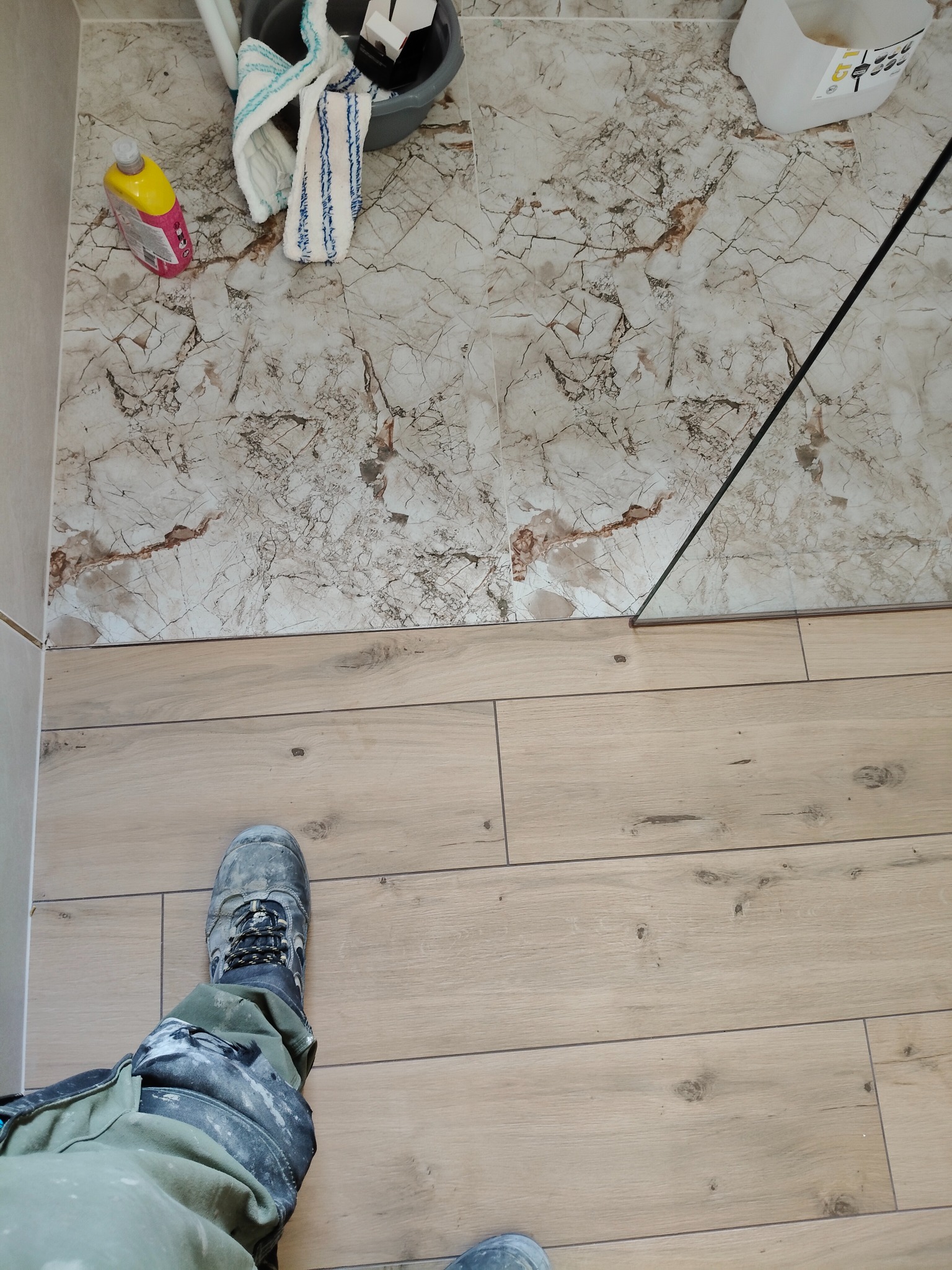 Ułożone płytki imitujące drewno na podłodze, z fragmentem marmurowych płytek na ścianie i narzędziami glazurniczymi. Widoczne buty i spodnie robocze glazurnika.